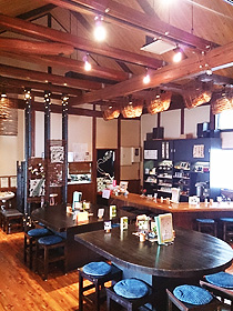 たくさんの梁が見える三角屋根の天井から電球が下がっており、楕円形のテーブル席やL字型のカウンター席がある「農家レストラン まだ来すた」の店内の写真