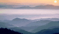 濃い霧の中、山々が連なっている奥で太陽が見えている幻想的な写真