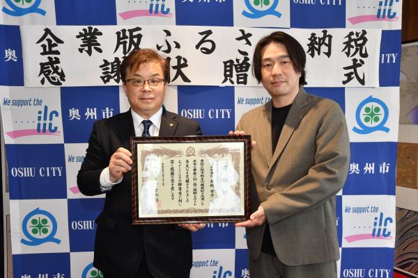 「企業版ふるさと納税感謝状贈呈式」の横断幕が掲げられた奥州市のバックボードの前で一緒に感謝状を持って立つ倉成市長と角地取締役社長の写真