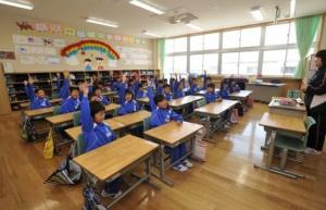 教室内にジャージ姿の子ども達が着席し、正面に立っている先生の方を向いて右手を挙げている写真