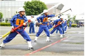 教官が見守る中、青い活動服にオレンジ色のゼッケンを身に着けた団員たちがホースを持ち放水訓練をしている写真