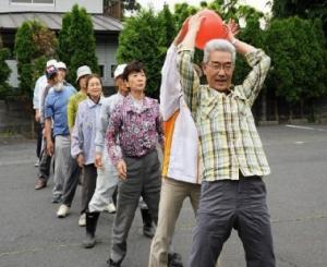 屋外で10名ほどの人が同じ方向を向いて縦一列に並び、先頭の男性から後ろの人へ赤いボールを正面を向いたまま両手で手渡している様子の写真