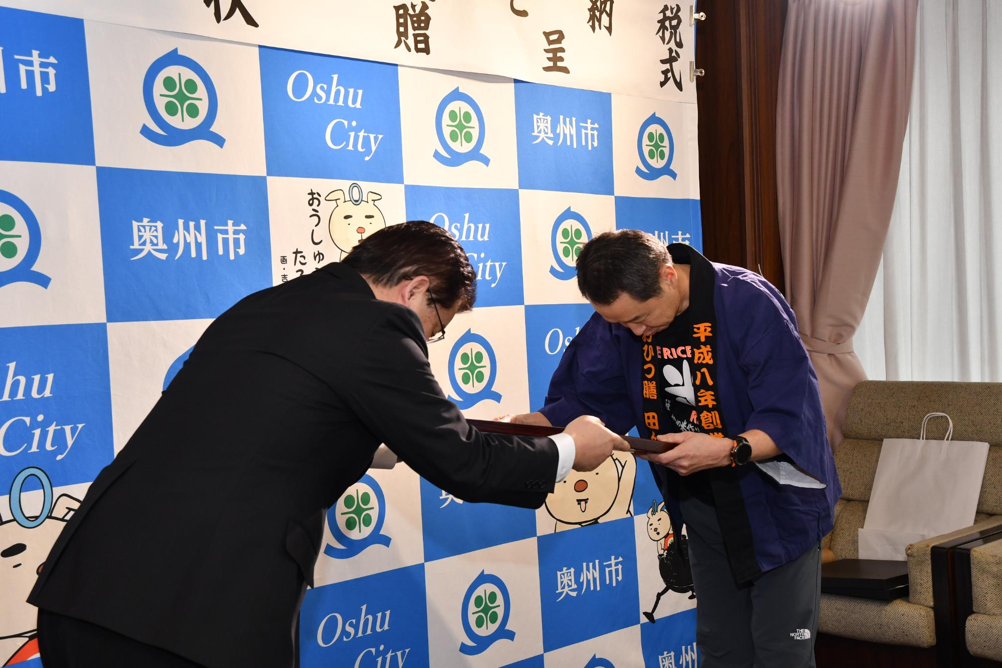 「企業版ふるさと納税感謝状贈呈式」の横断幕が掲げられた奥州市のバックボードの前で中川専務取締役に感謝状を手渡す倉成市長の写真