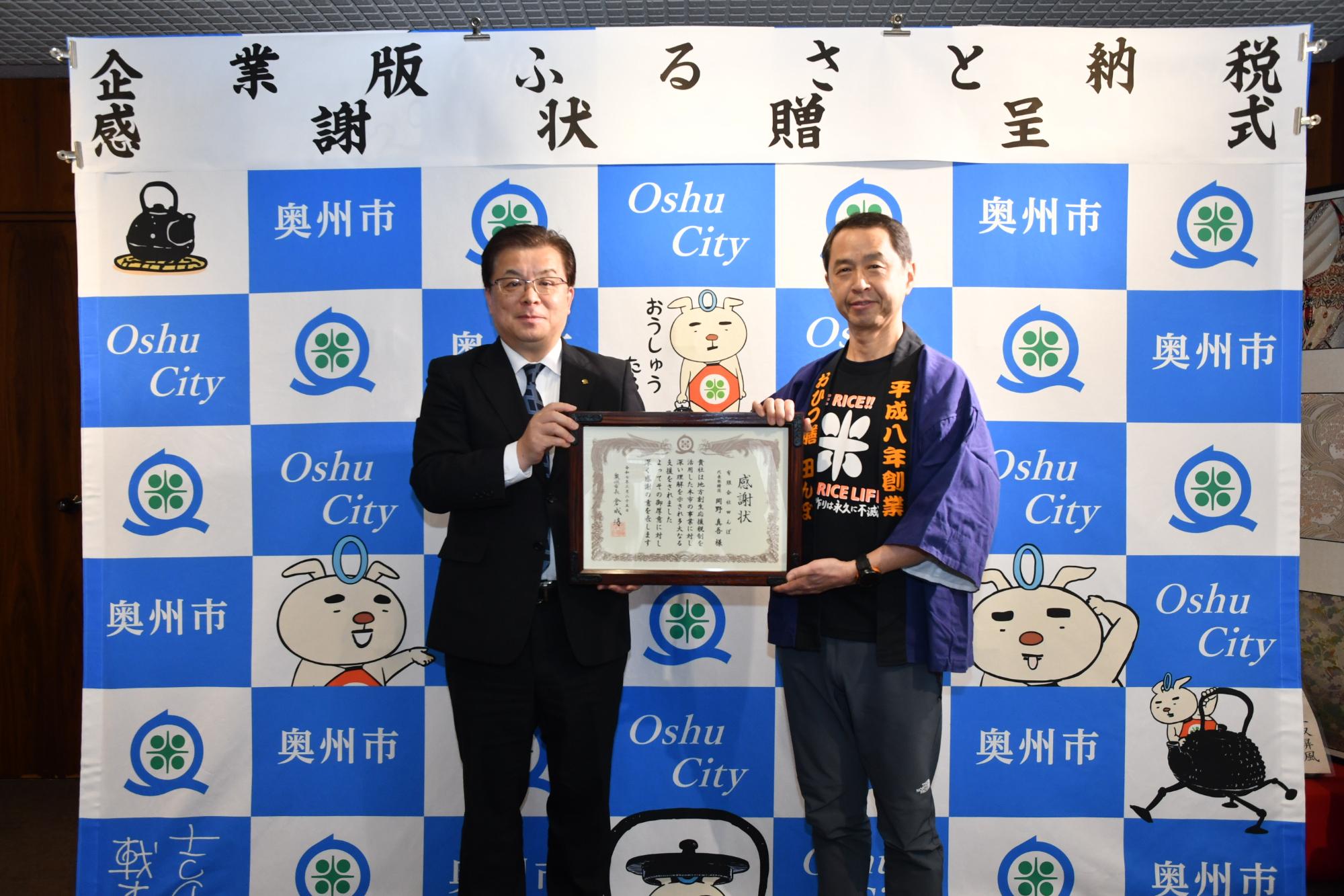 「企業版ふるさと納税感謝状贈呈式」の横断幕が掲げられた奥州市のバックボードの前で2人で感謝状を持って立つ倉成市長と中川専務取締役の写真