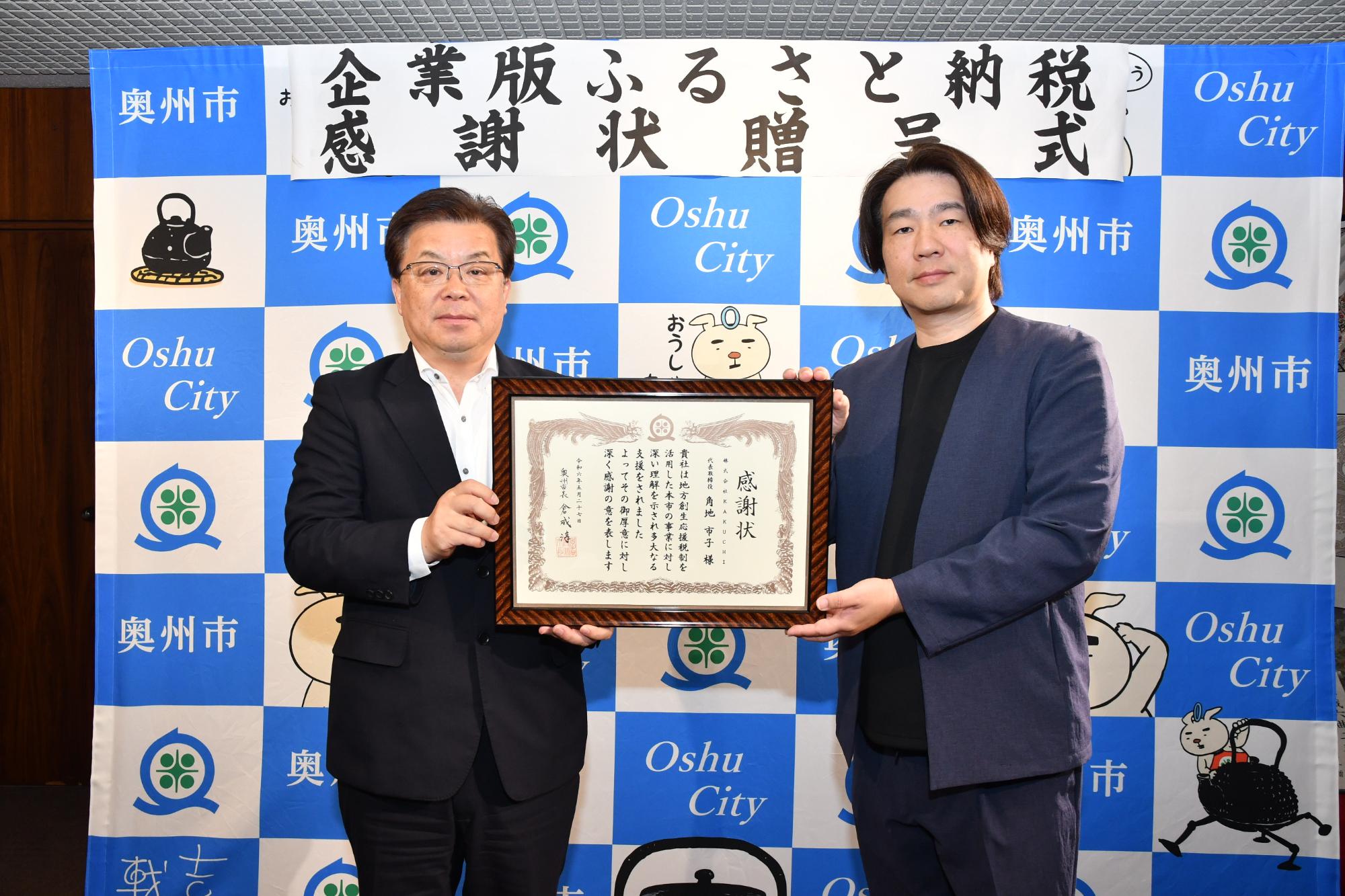 「企業版ふるさと納税感謝状贈呈式」の横断幕が掲げられた奥州市のバックボードの前で一緒に感謝状を持って立つ倉成市長と角地取締役社長の写真
