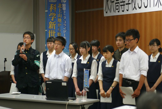 会場の前方に制服を着た11人の高校生が立ち、1人の男子高校生がマイクを持って話をしている写真