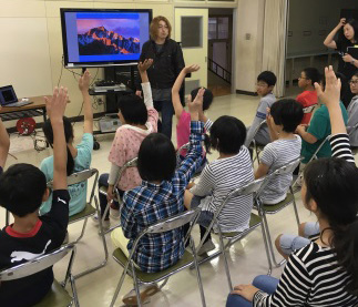 前方にモニターが設置された会場で齋藤武彦教授が立っており、子どもたちがパイプ椅子に座って手を上げている特別授業の様子の写真