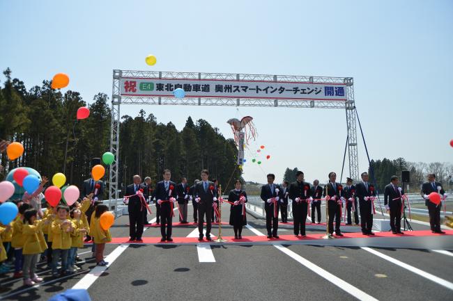 上に「祝E4東北自動車動奥州スマートエンターチェンジ開通」と書かれ、風船を持った園児達の横で関係者の方々がテープカットをしている写真