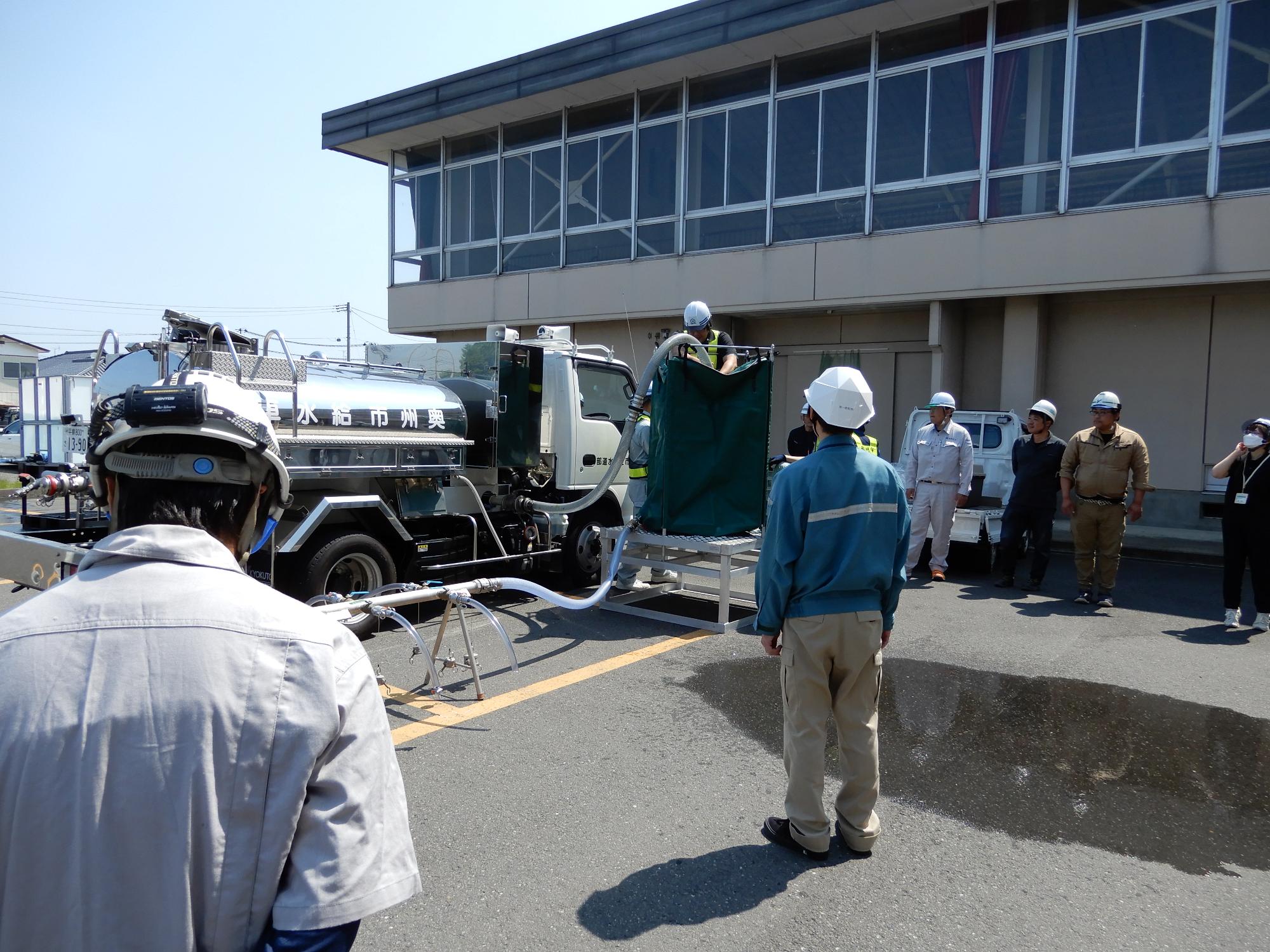 地面に設置された給水タンクへ給水車から補水を行う作業員と、それを見学する参加者の様子の写真