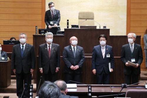 議場の演壇の前に5名の教育厚生常任委員会の方々が横並びで立ち、奥の議長席の横に男性が立っている写真