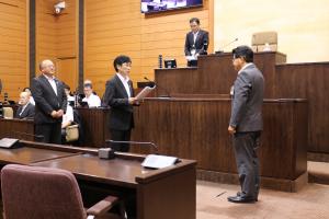 議場の演壇の前で、市長の前に2名の議員が立ち、代表して菅原由和総務常任委員会委員長が市長へ政策提言書を手渡す様子の写真
