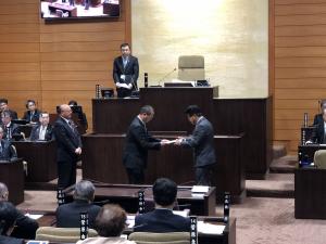 議場の演壇の前で、市長の前に3名の議員が立ち、代表して藤田慶則産業経済常任委員会委員長が市長へ政策提言書を手渡す様子の写真