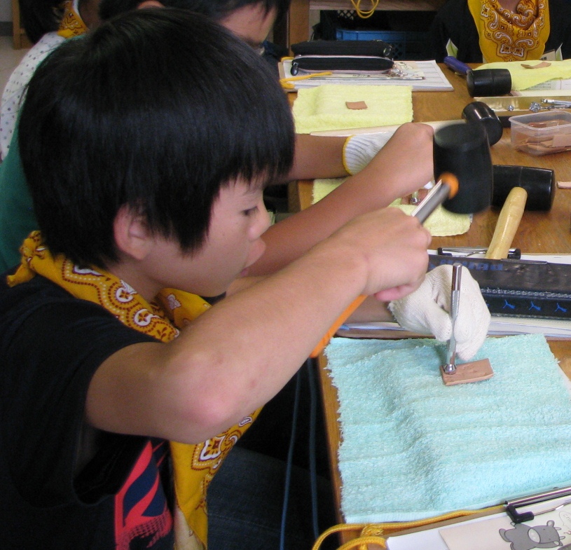 男の子がテーブルにタオルを広げ、トンカチを使って、長方形の革に丸い形を打ち付けている作業を横から写した写真