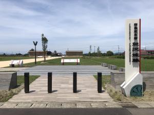 薄い雲が広がる中、手前の胆沢城跡歴史公園入口には、国指定史跡胆沢城跡と書かれた園名板と車が進入しないよう4本のポールが設置され、左側には政庁跡へと続く白い南大路が北へ真っ直ぐ続いている写真