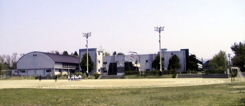 グラウンドで体操服姿の生徒たちが立っている先に、かまぼこ屋根の体育館や白い外壁の校舎が建っている奥州市立羽田小学校の外観写真