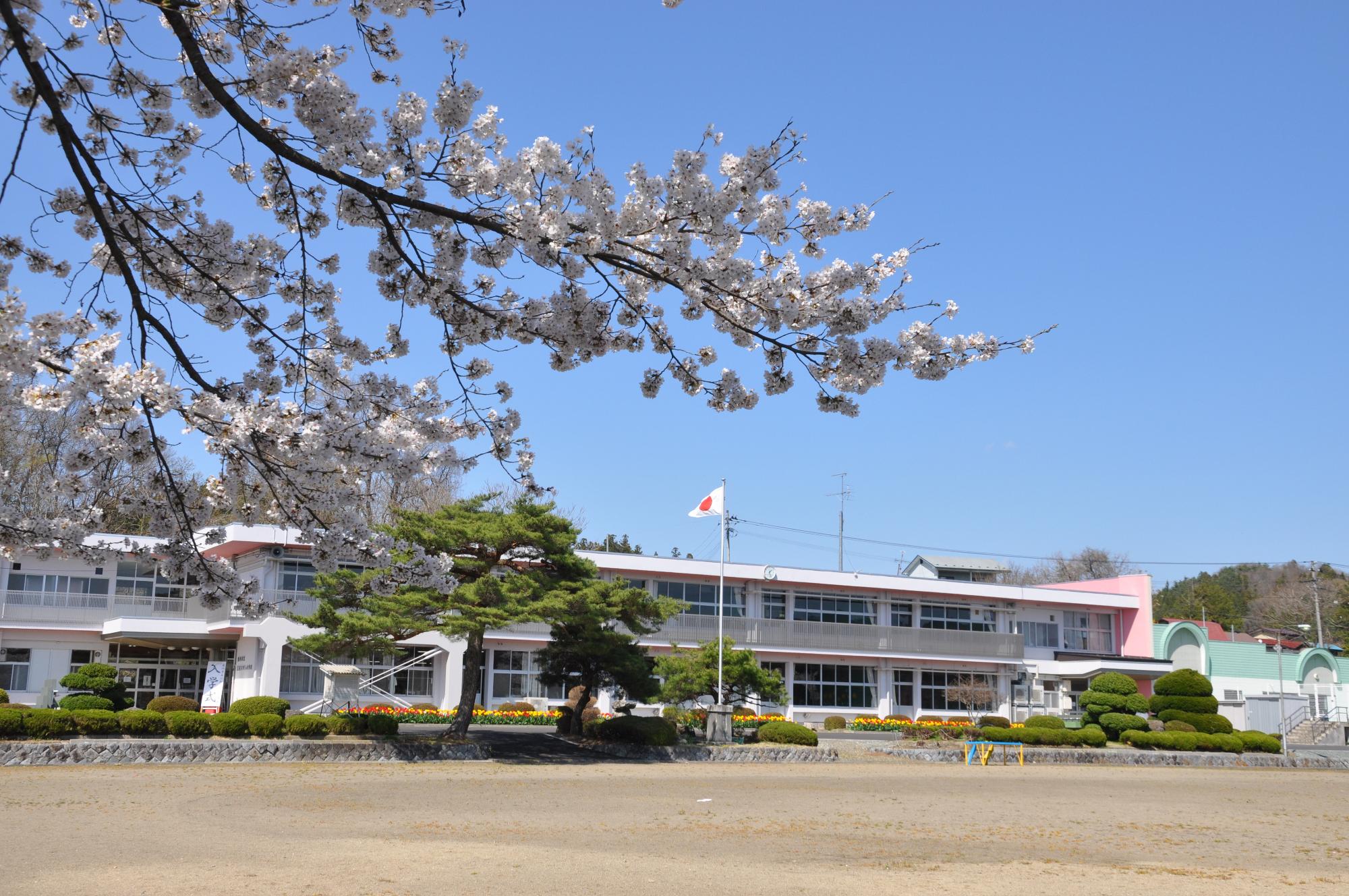 校舎の前には多くの植木があり、2階建ての校舎の先には林が広がっている。自然豊かな奥州市立江刺ひがし小学校の外観写真（春）